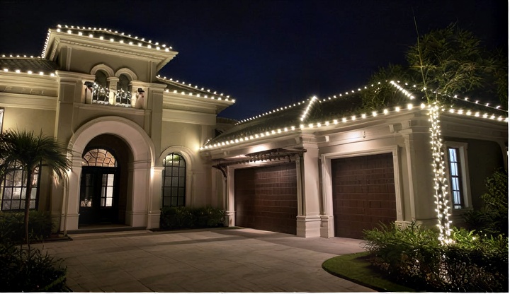 Large two-story Mediterranean-style Palm Harbor home at night with warm white string lights outlining the rooflines, eaves, arches, and a wrapped tree