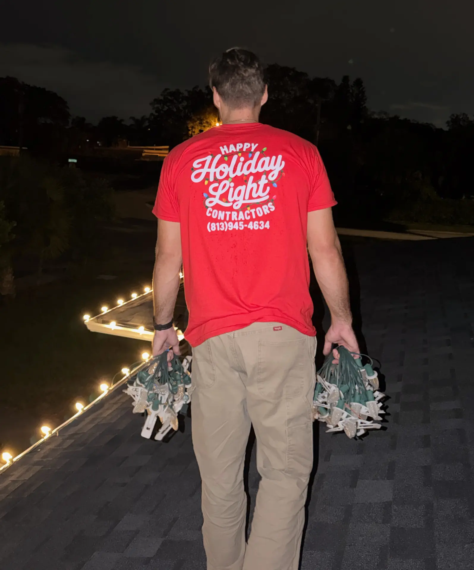 Happy Holiday Light Contractors crew member on a roof holding bundles of Christmas lights at night, wearing a red company shirt